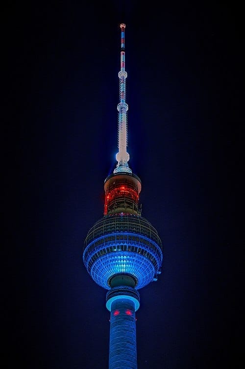 The top of the Berlin Television Tower. It is a thin metallic structure ending in a sphere and spire with red lights. There is black sky behind it.